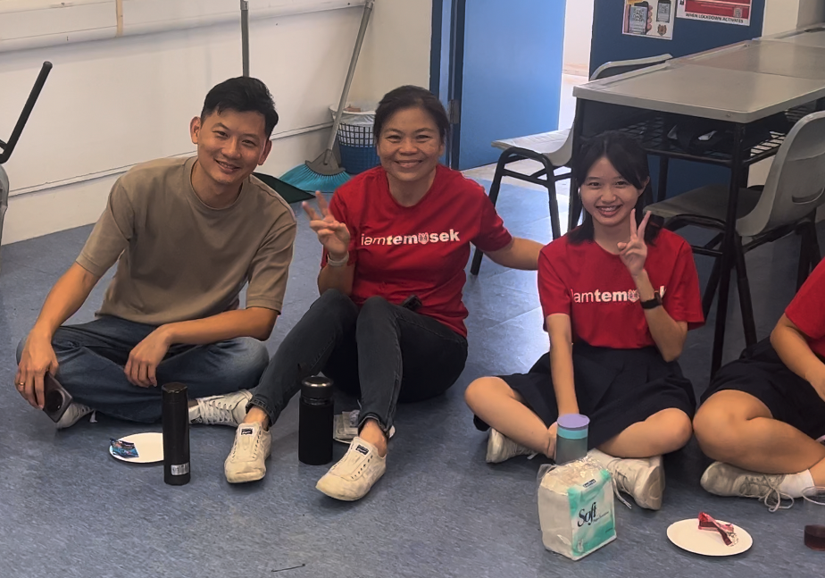 Three smiling people sit on a floor, two wearing red iamtemasek shirts, making peace signs.